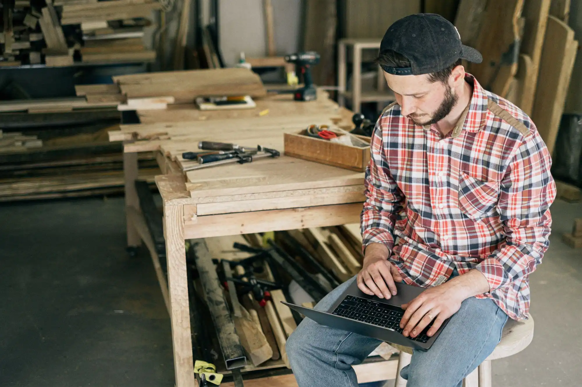 A tradesman in a plaid shirt working on a laptop in his woodshop